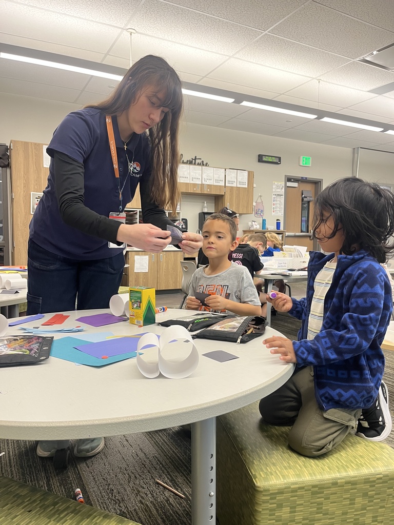 A female teacher or staff member stands next to two young students seated at a low circular table, helping them with a craft project involving paper loops and colored shapes. The children are engaged, with art supplies and paper scattered on the table. The classroom environment is bright and modern with visible learning materials.