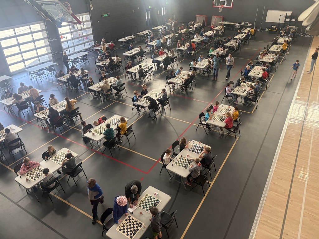 A large group of students sit at small tables playing chess during a tournament in a school gymnasium.
