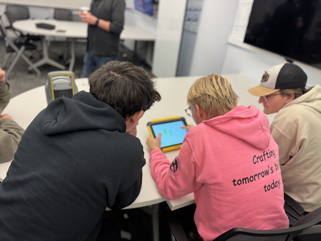 Three students sit at a table working with a tablet device in a tech lab. One student wears a black EPIC Campus hoodie, and another wears a bright pink hoodie that reads “Crafting tomorrow’s built world today.” A grey and yellow scanner device sits on the table nearby. A fourth person, holding a coffee cup, stands in the background.