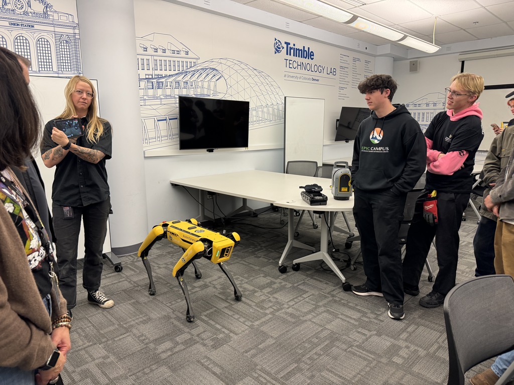 A group of students and a presenter stand around a yellow quadruped robot labeled “Spot” in the Trimble Technology Lab at the University of Colorado Denver. One student wears an EPIC Campus hoodie. The presenter, with long blond hair and visible tattoos, holds a tablet while explaining the robot. A line drawing of Union Station and other architecture is displayed on the wall in the background.