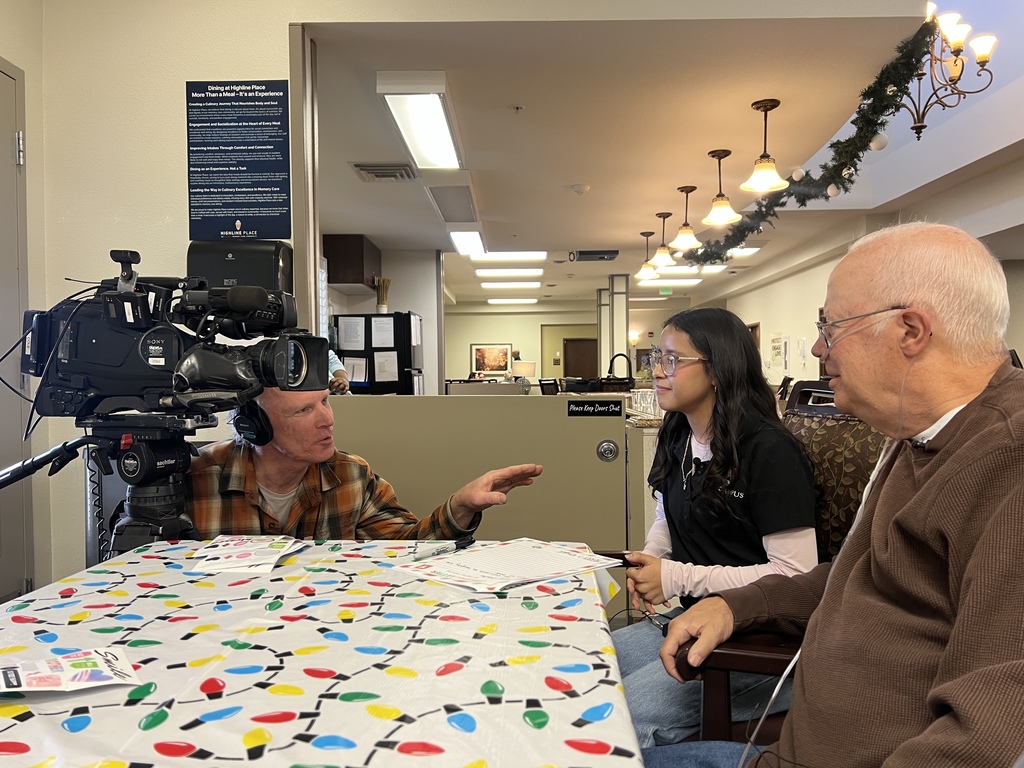 A student in an EPIC Campus shirt and an older man sit side by side at a holiday-decorated table, being interviewed on camera by a man operating professional video equipment. The student is mid-conversation, smiling, while the cameraman gestures toward her. The setting is a senior living facility with warm lighting and holiday garland overhead.