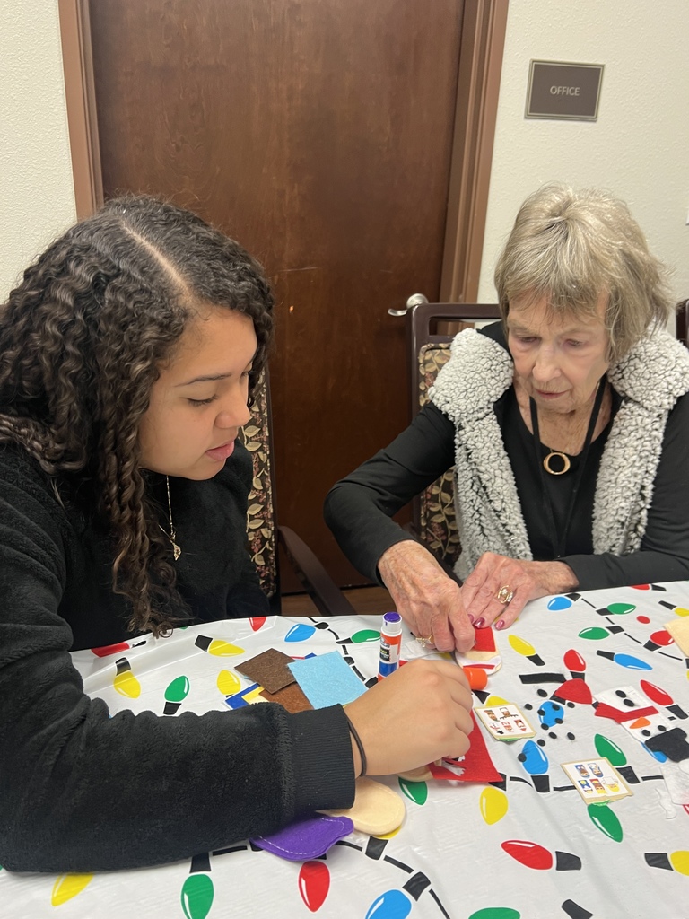A student and an elderly woman work on a holiday-themed craft project together at a table covered in a string light print tablecloth. The table is scattered with felt, glue sticks, and colorful shapes. Both are focused on gluing pieces to create ornaments or decorations. A brown door with an “Office” sign is in the background.