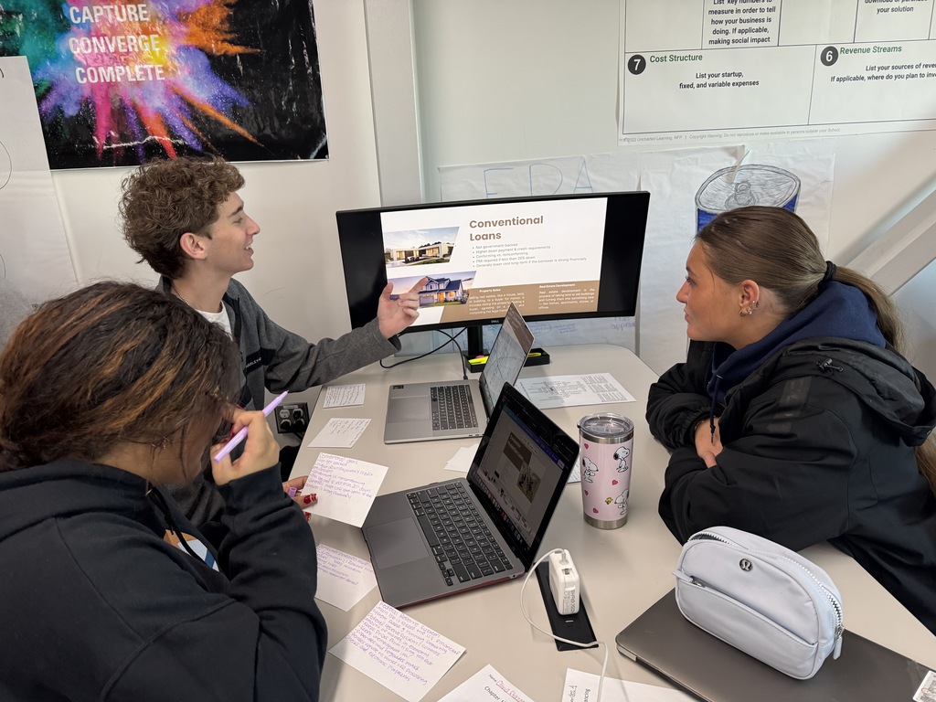 Three students collaborate around a table with laptops and notecards while presenting a slide titled "Conventional Loans" on a monitor. One student gestures toward the screen while the others listen. A pink Snoopy tumbler and white pencil pouch are on the table. Informational posters and charts are visible on the walls around them.