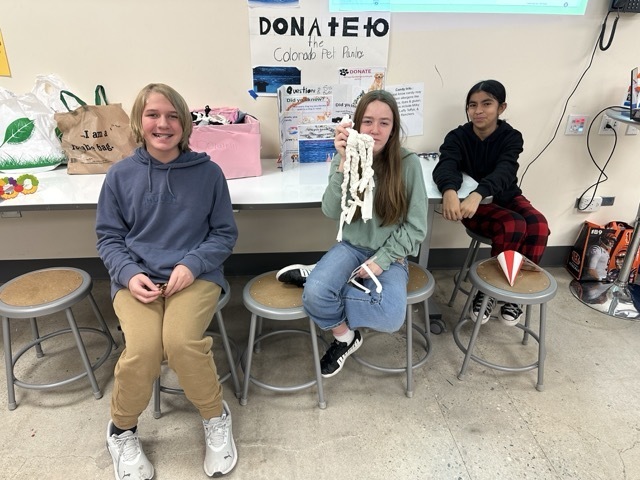 Three students sit at a table with a "Donate to the Colorado Pet Pantry" sign and handmade dog toys.