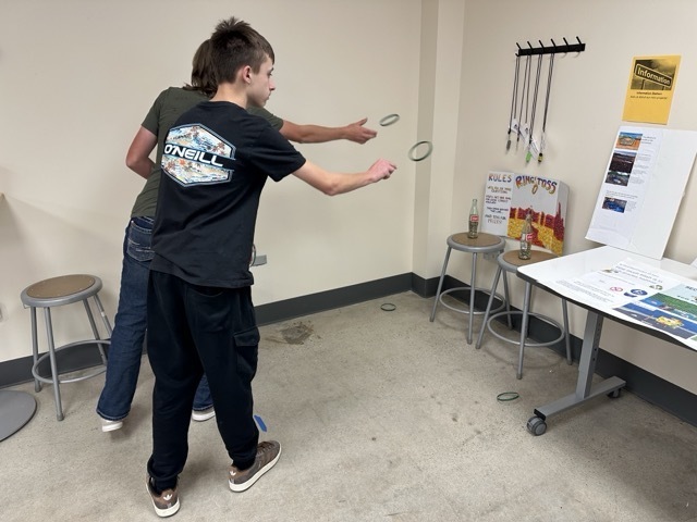 Two students toss green rings toward glass bottles in a homemade "Ring Toss" carnival-style game.