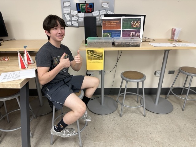 A student gives a thumbs-up while sitting next to a science project about how compost affects soil.