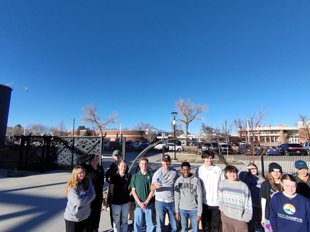 A large group of students and adults pose for a photo in front of a school building on a bright, sunny day. They stand on a sidewalk near a metal gate with decorative cutouts, and several students wear EPIC Campus shirts or sweatshirts. Behind them are leafless trees, parked cars, and school buildings. The mood is cheerful and collaborative.
