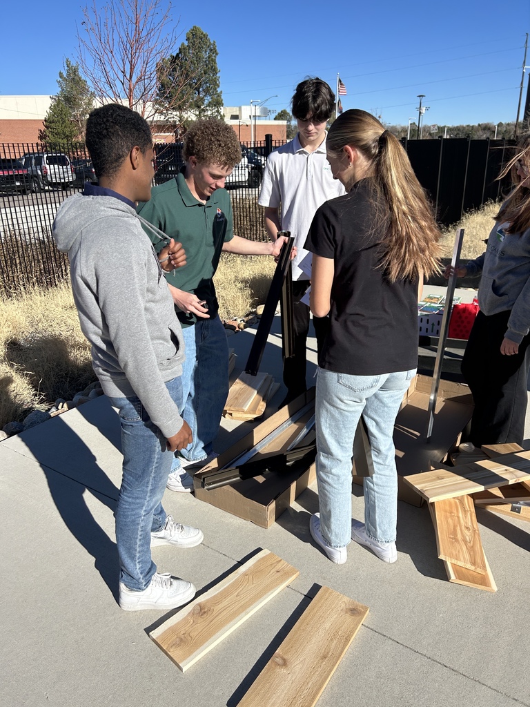 A group of high school students outdoors assembling a curved metal grid structure on a paved walkway. One student crouches, attaching parts at the base while others stand nearby, observing. The students wear casual clothing and EPIC Campus gear. In the background are leafless trees, dried landscaping, and an orange metal pergola.