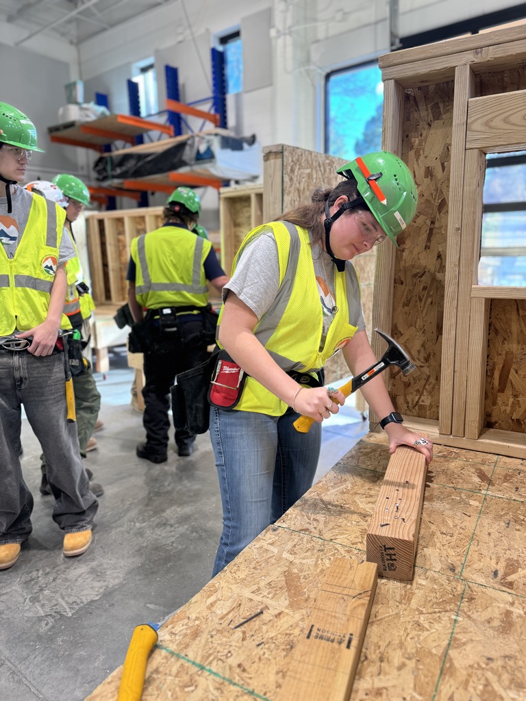 Two students in safety gear work together in a construction lab. One student stands on a platform holding a vertical wooden beam while the other, wearing a “Brave” stickered hard hat, aligns it from below. Both wear green hard hats, yellow safety vests, and tool belts. Nearby students observe.