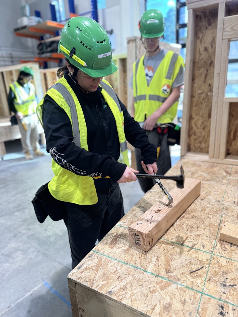 A group of students in green hard hats and yellow vests work around a raised wooden platform, with one student hammering a nail into a piece of wood while others observe. A teacher or mentor in a white hard hat and tattooed arm stands nearby, guiding the process. The setting is a bright, well-equipped construction classroom.