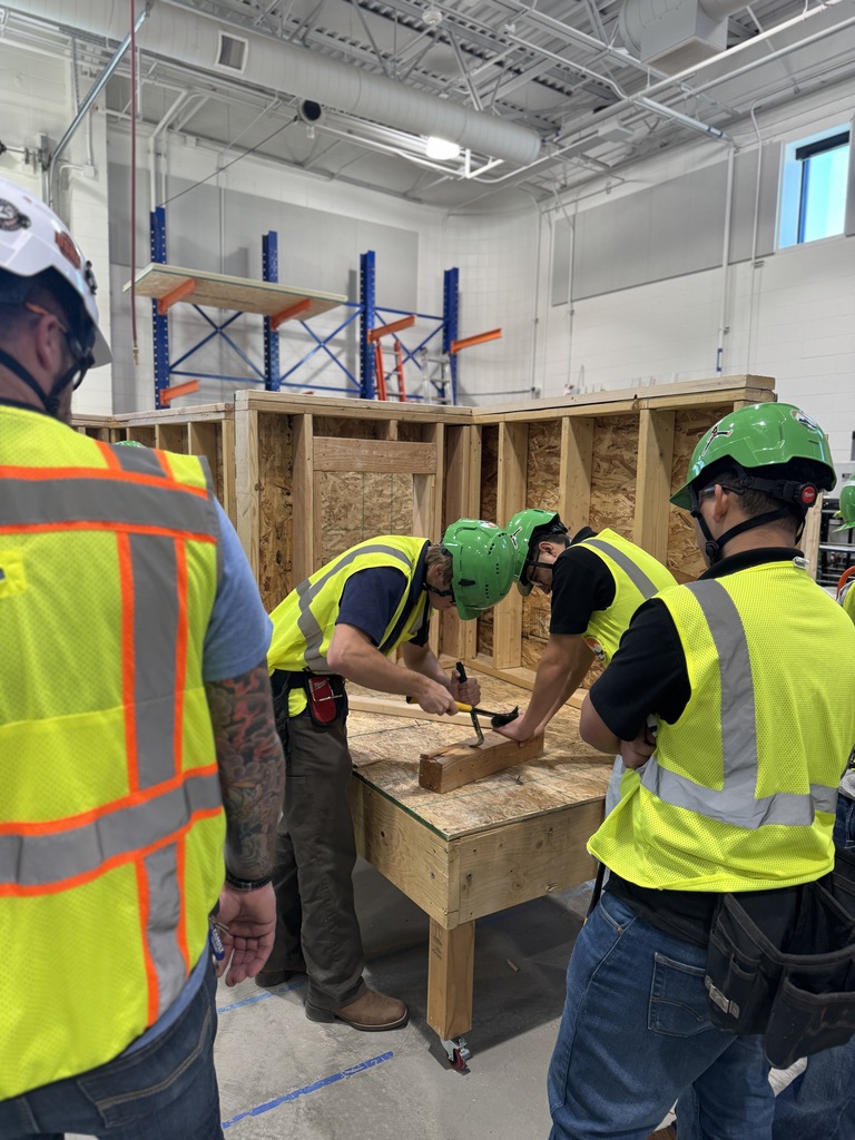 A student in a green hard hat and yellow vest uses a hammer to drive nails into a wooden block on a workbench. Another student stands behind her observing. The room contains multiple wood-framed structures in progress and shelves with tools and materials.