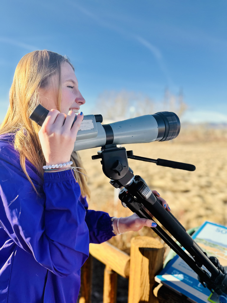 A student in a purple jacket smiles while using a spotting scope mounted on a tripod during an outdoor field study. A blue sky and dry grassy landscape stretch behind her.