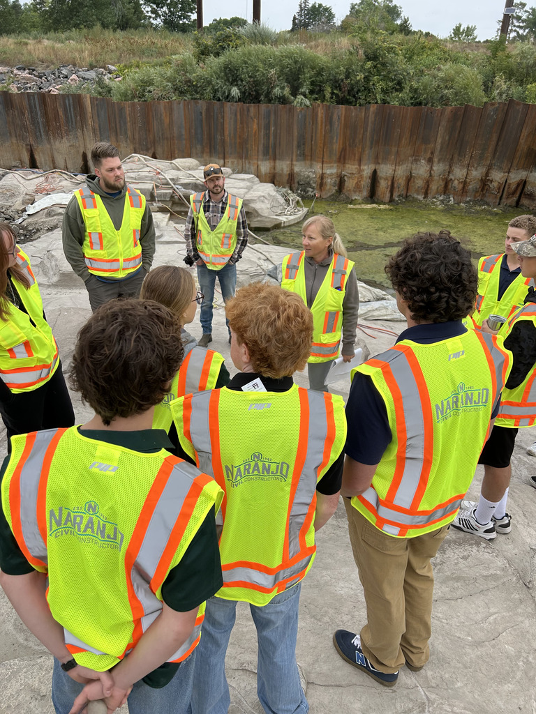 Students and adults wearing neon yellow safety vests with "Naranjo Civil Constructors" branding stand in a construction zone outdoors. They listen to a speaker while surrounded by heavy-duty barriers and vegetation.