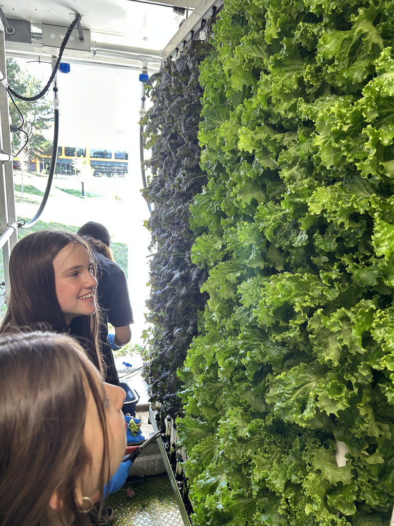 Students work inside a hydroponic farm trailer, smiling as they inspect vertical rows of lush green and purple lettuce. The tightly packed greens are growing along metal walls under artificial lighting.