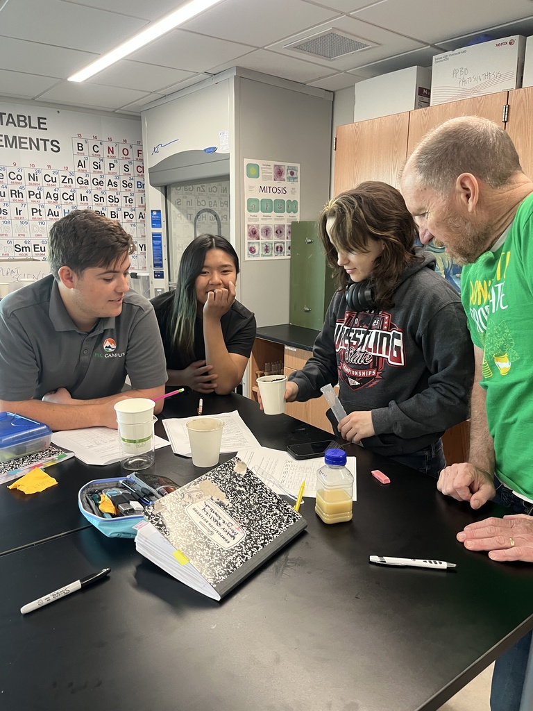 Four students and a teacher gather around a black science lab table in a classroom. They examine small cups and a pipette, with notebooks, a composition book, and science posters visible in the background.