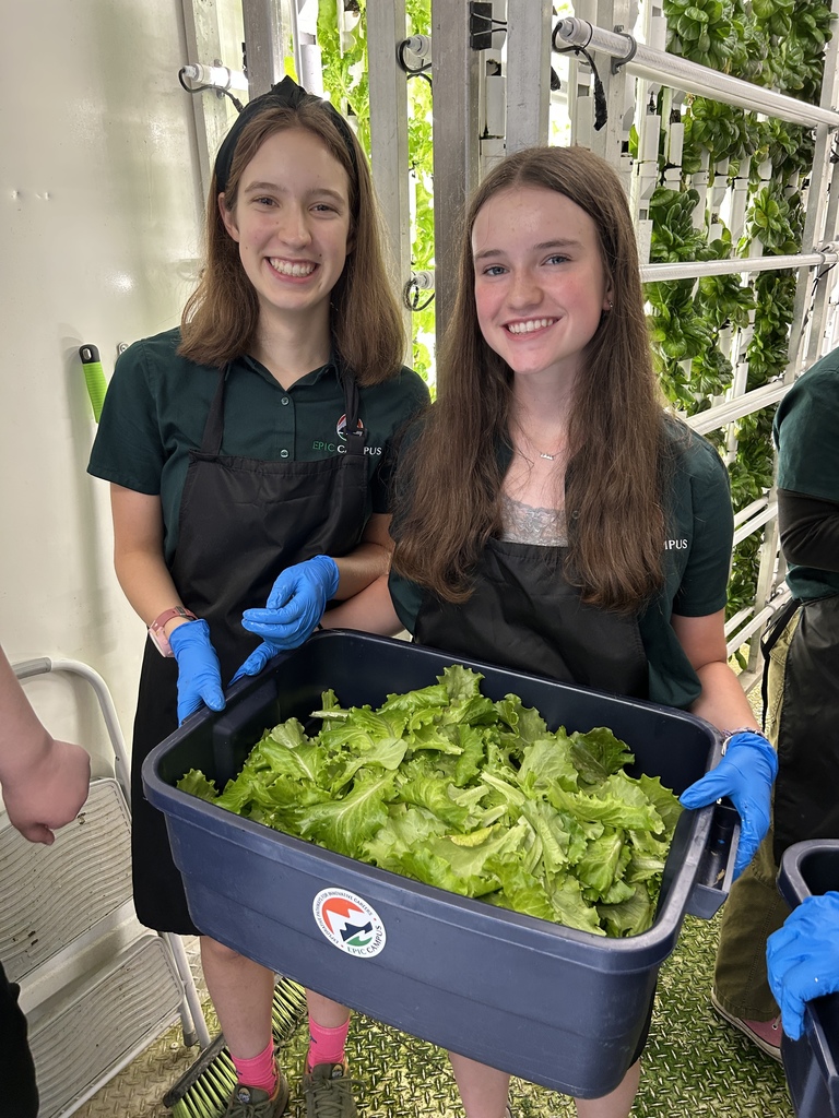 Two smiling students wearing EPIC Campus uniforms and aprons hold a large tub filled with freshly harvested lettuce inside a hydroponic growing facility. Vertical rows of leafy greens fill the background.