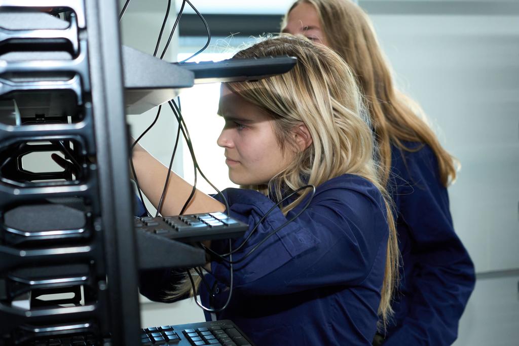 Two students in dark blue shirts work with tangled computer wires in a tech lab. One student is closely examining and connecting cables on a multi-level metal rack of monitors and keyboards.