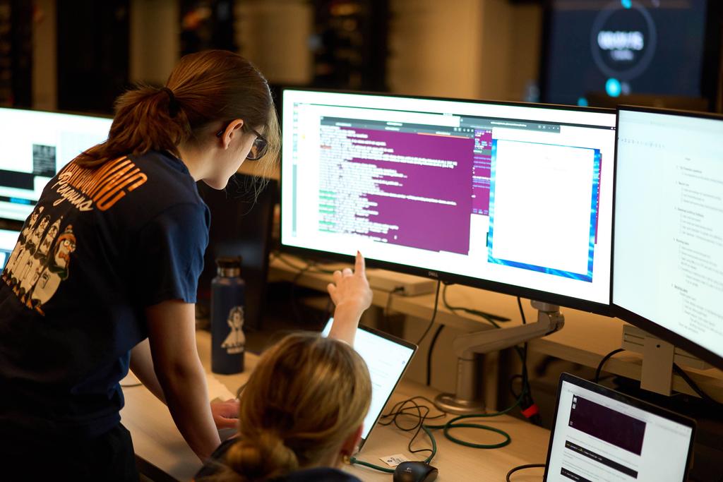 Two students focus on lines of code on large computer monitors in a dimly lit lab. One student points at the screen while the other leans over a laptop. A shirt design reading “Programming Penguins” is visible.