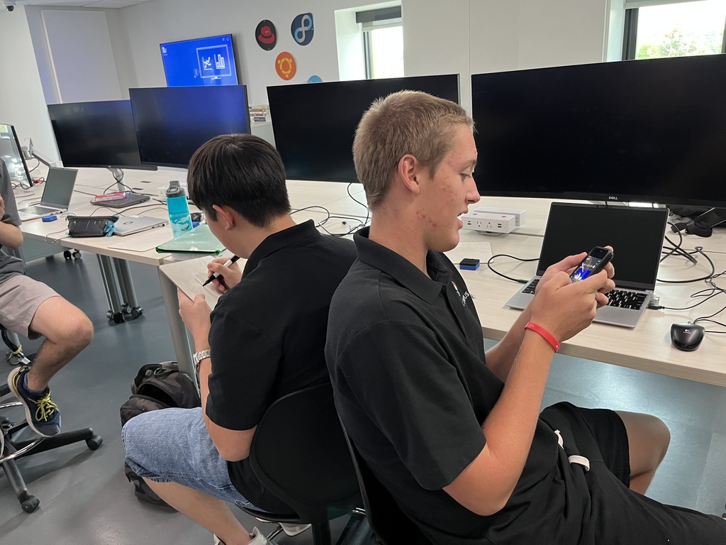 Two students sit back-to-back at a tech classroom table, each interacting with handheld devices. The room features laptops, large monitors, and programming-related wall logos.