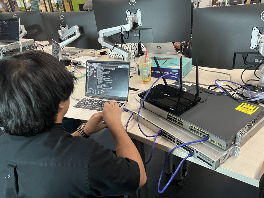 A student works on coding at a laptop connected to a wireless router and stacked network switches. Multiple desktop monitors and tech equipment fill the classroom.