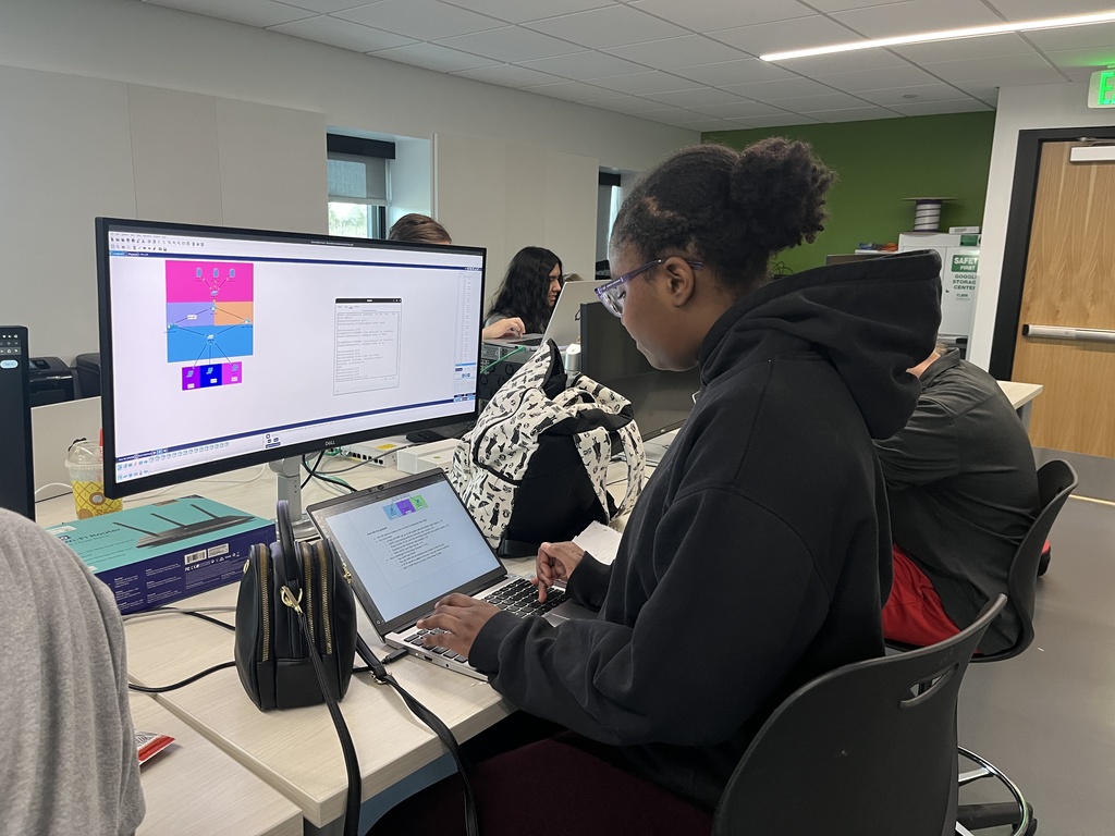 A student in a black hoodie types on a laptop connected to a desktop monitor displaying a colorful network diagram. The workspace includes tech boxes and a small handbag, with other students in the background.