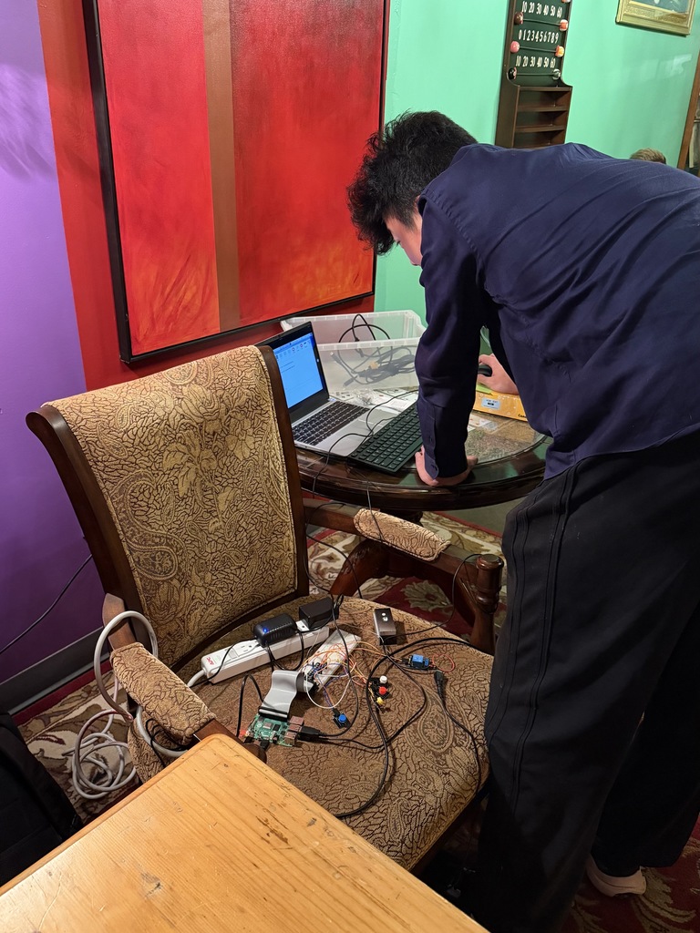 A student leans over a laptop connected to wires and electronic components on a chair. The setup includes a power strip, Raspberry Pi board, and various buttons. The student works in a colorfully painted room with red, purple, and green walls.
