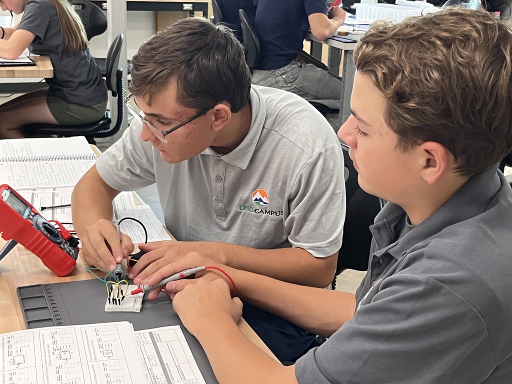 Two male students in gray EPIC Campus polos work together on an electronics project, using a breadboard and multimeter. They are seated at a desk covered in circuit diagrams and class materials.
