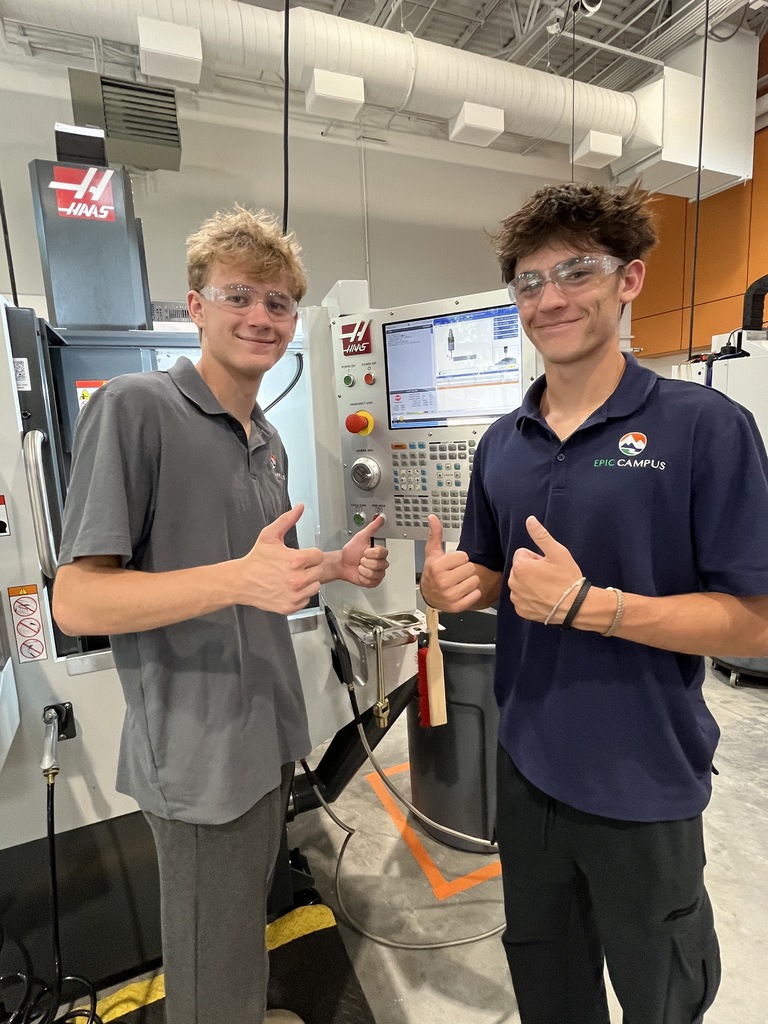 Two male students wearing EPIC Campus polos and safety glasses stand in front of a HAAS CNC machine, smiling and giving thumbs up. The background shows a clean, well-lit workshop.