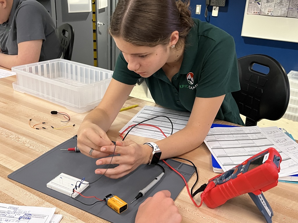 A female student in a green EPIC Campus polo carefully connects wires to a breadboard. She is focused on the task, surrounded by lab materials, circuit sheets, and a red multimeter.