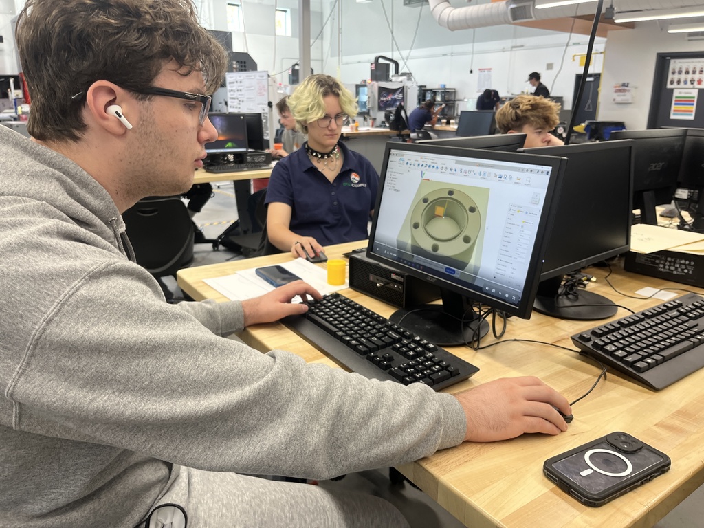 Two students work at desktop computers using CAD software to design a mechanical part. One wears a gray hoodie and AirPods; the other wears a navy EPIC Campus shirt and choker necklace. Their monitors show a 3D model.