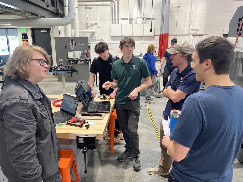 A group of six high school students, mostly male and wearing EPIC Campus shirts, stand around a workbench in a modern manufacturing lab. Two open laptops sit on the bench alongside tools and red cables. One student in a green polo appears to be explaining something, while others listen or observe. The workspace includes industrial machines and bright lighting, with additional students working in the background.