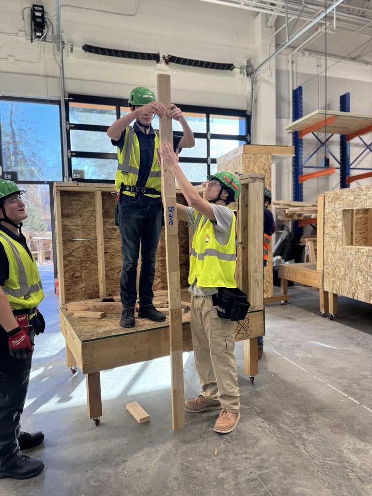 One student stands on a wooden platform while another steadies a tall vertical beam marked "Brave." Both wear green hard hats, safety vests, and tool belts. Other students and construction materials fill the background of the bright workshop.