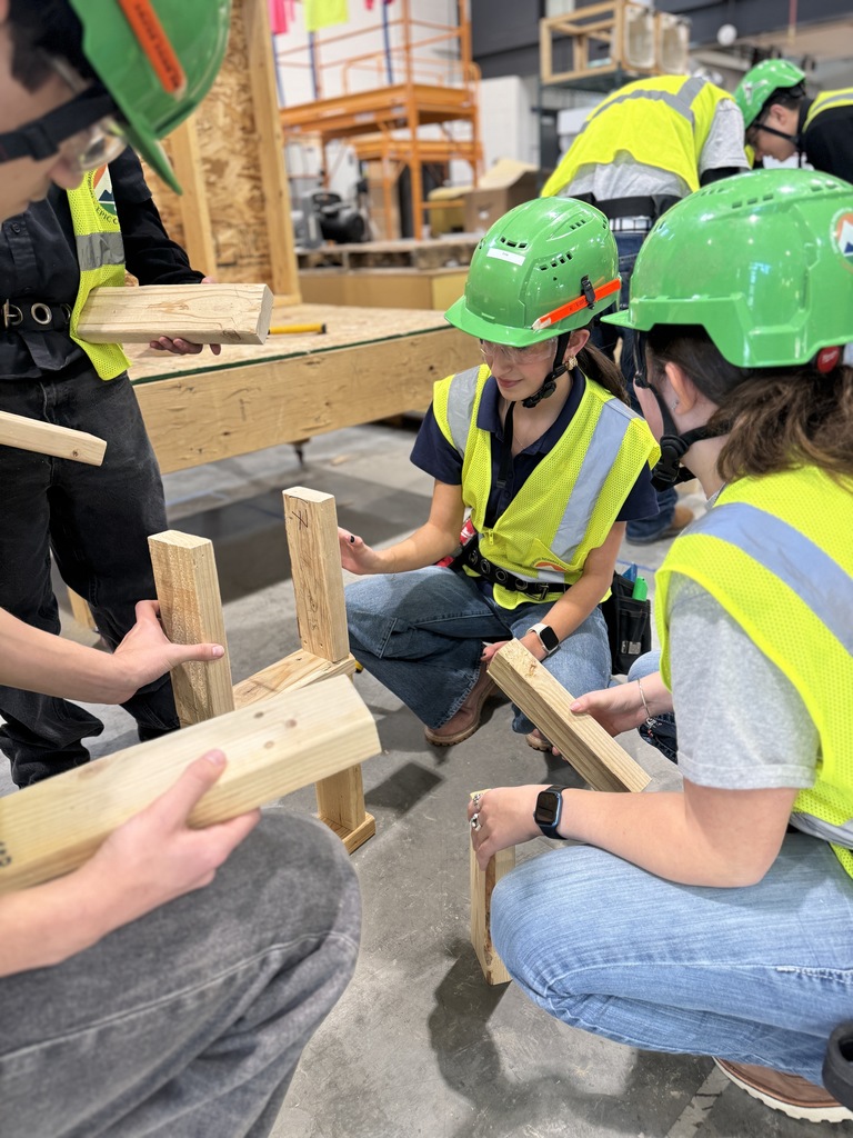A group of students in green hard hats and yellow safety vests kneel and stand in a circle, collaborating to assemble a wooden structure using small cut pieces of lumber. They are engaged in discussion and hands-on work in a construction lab filled with tools and workbenches.