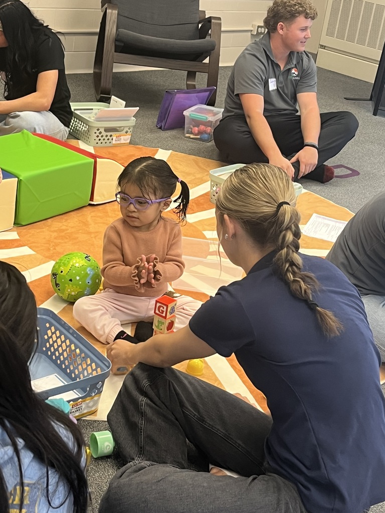 A young girl with glasses sits on a sun-themed carpet, playing with blocks and toys while interacting with a high school student in an EPIC Campus shirt. Other students and children are also engaged in play around the room.