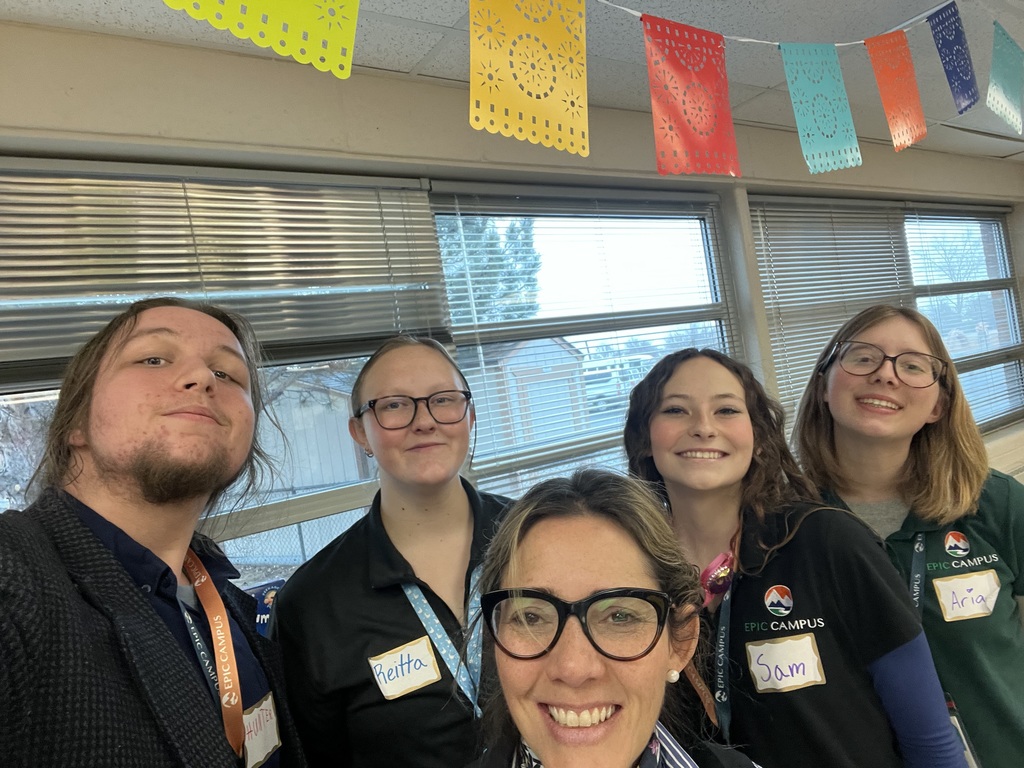 A group selfie featuring four high school students and one adult, all smiling in a classroom. Three students wear EPIC Campus shirts and name tags, and colorful papel picado decorations hang above them. Windows line the background wall, letting in daylight.