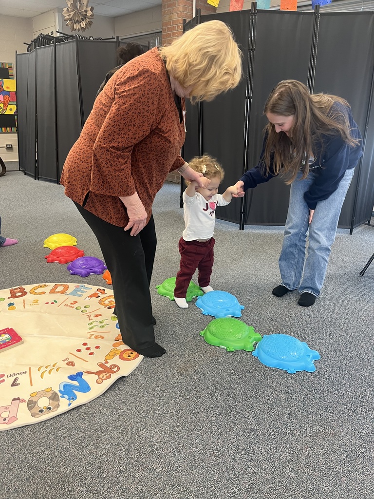 A toddler walks across colorful stepping stones with the help of an older adult and a high school student. An alphabet-themed rug and various learning toys are visible in the classroom, which is well-lit and cheerful.