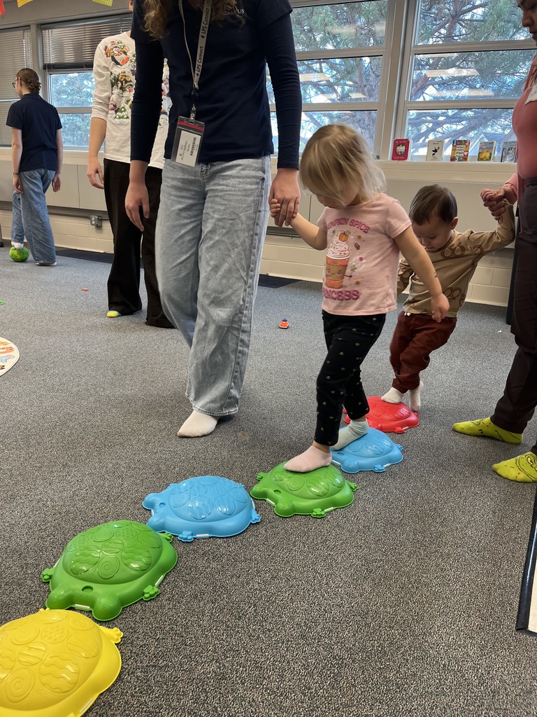 Two young children walk barefoot across a path of colorful turtle-shaped stepping stones, guided by adults holding their hands. High school students in EPIC Campus shirts are visible in the background, assisting in the activity inside a bright classroom with large windows.