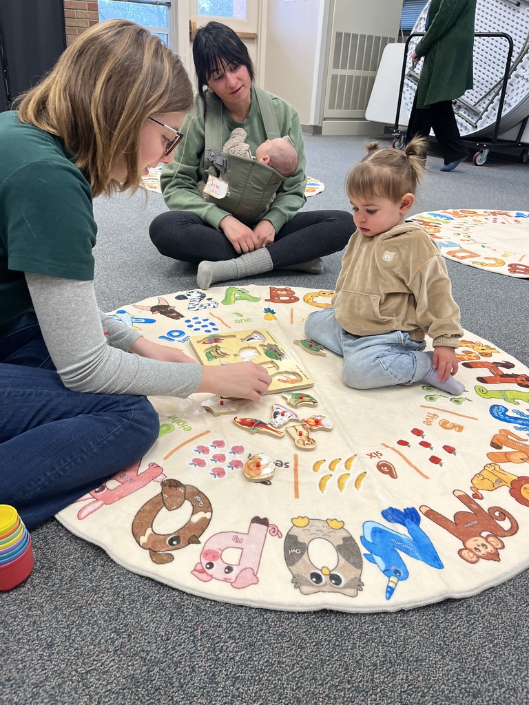 A high school student works on a puzzle with a toddler on an alphabet-themed rug, while a seated adult nearby holds a baby in a front-facing carrier. The room is filled with educational mats and playful learning materials.