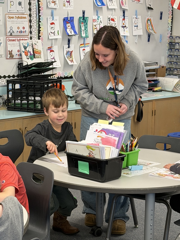 A high school student in an EPIC Campus hoodie leans over to help a young boy with his worksheet at a classroom table. They are both smiling. The background displays colorful student artwork and learning materials.