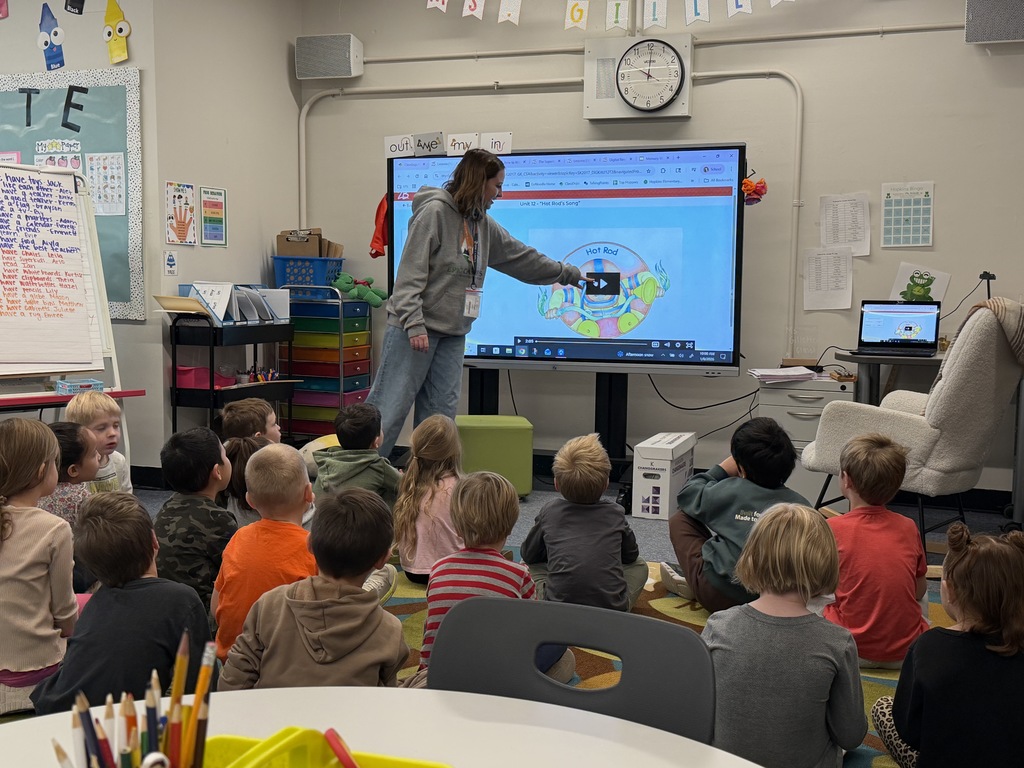 A high school student in an EPIC Campus hoodie stands at the front of a classroom, pointing to a large interactive screen displaying a video titled “Hot Rod’s Song.” Around 20 young children sit cross-legged on the carpet, attentively watching the screen. The classroom is colorful and well-organized, with bins, posters, and student work on display.