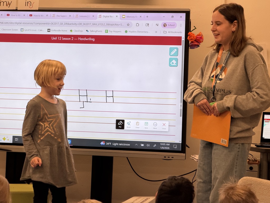 A young girl stands in front of the class smiling, while a high school student in an EPIC Campus hoodie stands nearby, also smiling. Behind them, a large screen displays a handwriting lesson titled “Unit 12 Lesson 2 – Handwriting” with lines and letters drawn on a digital whiteboard.