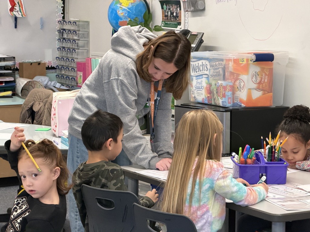 A high school student in an EPIC Campus hoodie bends over to assist a group of young students at a table. The children are working with pencils and markers on worksheets. The classroom is colorful and organized, with a globe, snack boxes, and learning supplies visible.