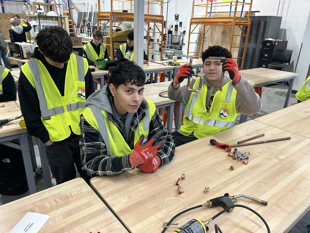 Two students at a workbench, one holding up a completed copper pipe assembly while the other poses with a peace sign. Both wear yellow EPIC Campus safety vests and gloves. Their table is covered with plumbing tools, fittings, and pipe segments, and other students work in the background.