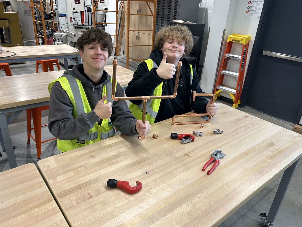 Two high school students wearing safety vests sit at a wooden workbench, proudly holding up their assembled copper pipe project and giving thumbs up. Various plumbing tools and fittings are scattered on the table around them in a bright, well-equipped workshop.