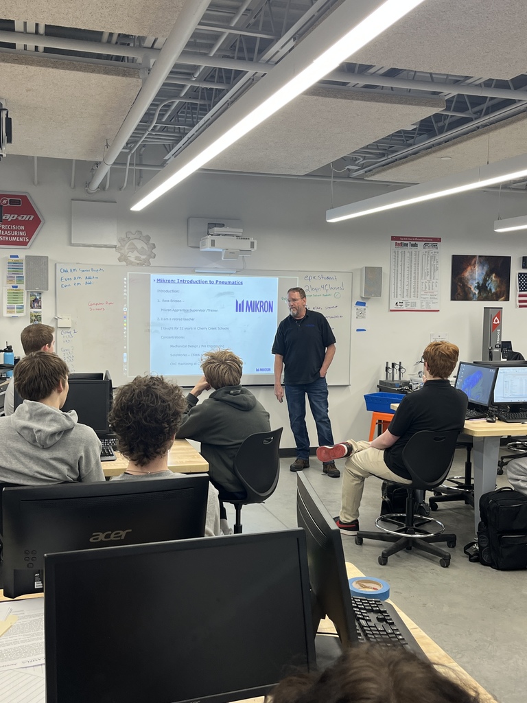 A male instructor stands in front of a large projected screen displaying a slide titled “Mikron: Introduction to Pneumatics.” Several high school students sit at desks with computers, facing the instructor in a modern classroom with whiteboards, posters, and a ceiling-mounted projector.