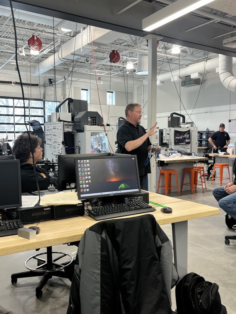 A male instructor gestures while speaking to a seated group of high school students in a large, open workshop. The space includes computer workstations and industrial equipment, with visible machines and storage areas in the background.