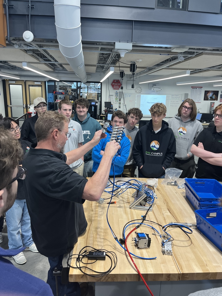 A male instructor demonstrates a pneumatic system with blue tubing to a group of attentive high school students gathered around a workbench. The students wear EPIC Campus apparel, and various components and tools are laid out on the table in a well-equipped lab.