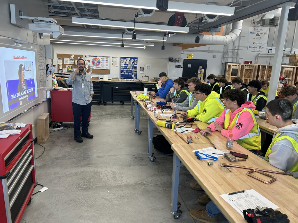 A male instructor stands at the front of a workshop classroom, gesturing as he addresses a large group of high school students wearing yellow safety vests. The students are seated at long workbenches with copper piping tools and materials in front of them. A motivational slide is projected on the wall behind the instructor, showing a speaker and the words "Handle Hard Better – Kara Lawson." The room is well-lit and filled with construction and workshop equipment.
