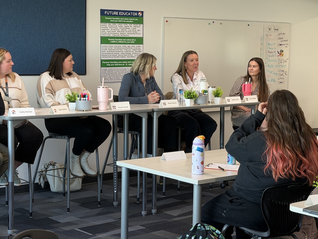 A close-up view of five women panelists, including educators and professionals, speaking and listening during a Future Educator panel. Students in the foreground observe attentively, and a whiteboard and posters are visible in the background.