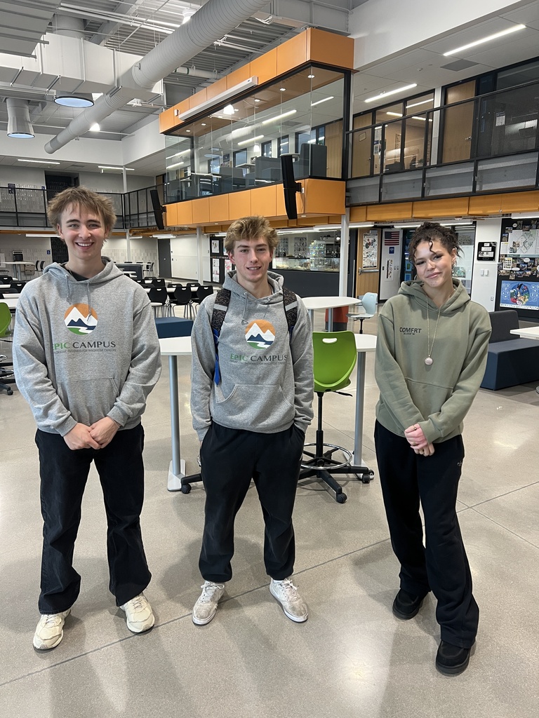 Three high school students standing in an open common area of a school building, wearing hoodies and sweatshirts. The background shows modern architecture with glass walls and orange accents.